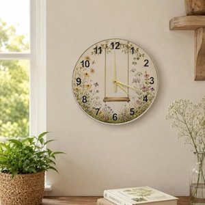 May include: A round wall clock featuring a cream-colored face decorated with a printed illustration of a wooden swing hanging from leafy branches surrounded by colorful wildflowers. The clock has black numerals and thin yellow clock hands.