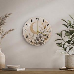 May include: A round wall clock with a cream-colored face featuring a gold crescent moon and botanical illustrations of leaves in shades of brown and gray. The clock has black numerals from 1 to 12 and thin gold-colored hour, minute, and second hands.
