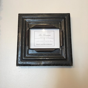 May include: A square, dark-toned picture frame with a layered design. The frame has a distressed finish and holds a white card with the text "One of a Kind Designs Tin Treasures Handmade Naomi and David Atkinson."