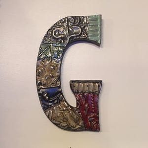May include: A decorative metal letter "G" with a detailed, embossed design. The letter features a mix of colors, including silver, blue, green, and red, with intricate patterns and textures. The letter is mounted on a white surface.