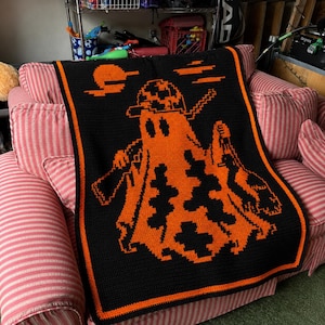 May include: A rectangular crochet blanket featuring a black background with a vibrant orange pixelated graphic of a ghost wearing a cap and holding a rifle. The design includes an orange sun and clouds, finished with a solid orange border trim.