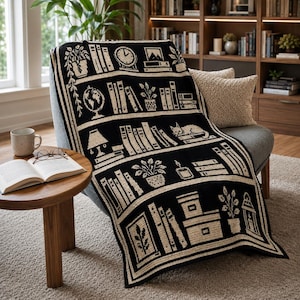 May include: A black and cream knit throw blanket featuring a repeating bookshelf pattern. The design includes illustrations of books, potted plants, a globe, a clock, a lamp, a sleeping cat, and boxes arranged on horizontal shelves with a border.