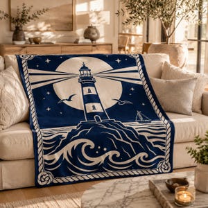Könnte beinhalten: Eine marineblaue und cremefarbene Wohndecke mit einer zentralen Leuchtturm-Grafik auf einer felsigen Klippe, umgeben von einem großen Mond, Sternen und fliegenden Vögeln. Das Design umfasst ein Segelboot auf dem Wasser und eine dicke Seilbordüre.