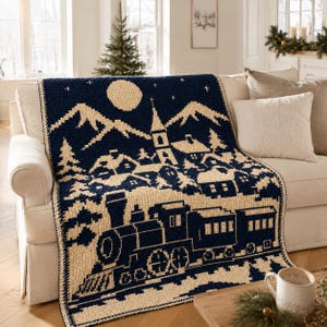 May include: A navy blue and cream crochet blanket featuring a winter village scene with a steam train in the foreground, a church steeple, snow-covered houses, mountains, and a full moon in the night sky, all rendered in a detailed tapestry pattern.