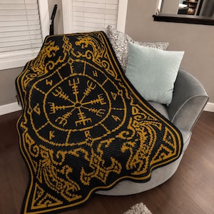 Op de afbeelding: Een zwart met gouden gehaakte deken met een centraal Vegvisir-symbool omringd door een cirkelvormige rand van runentekens. De hoeken en randen zijn versierd met ingewikkelde, symmetrische knooppatronen in contrasterend goudkleurig garen.