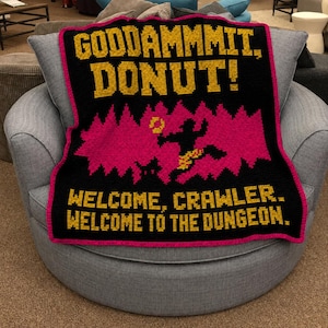 May include: A black crochet blanket with a pink jagged graphic in the centre featuring a silhouette figure. The text reads GODDAMMMIT, DONUT! at the top and WELCOME, CRAWLER. WELCOME TO THE DUNGEON. at the bottom in yellow block lettering.