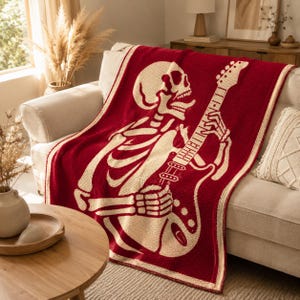 Könnte beinhalten: Eine rechteckige Strickdecke mit tiefrotem Hintergrund und einer großen, cremefarbenen Grafik eines Skeletts, das eine E-Gitarre spielt. Die Decke hat einen dicken, cremefarbenen Rand und eine durchgehend texturierte, gewebte Optik.