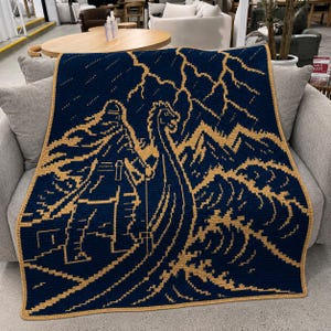 Puede incluir: Una manta de ganchillo rectangular con un fondo azul marino oscuro y un diseño gráfico en amarillo dorado que contrasta. El patrón muestra un barco vikingo estilizado navegando sobre olas turbulentas hacia montañas escarpadas.