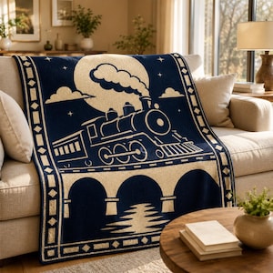 May include: A navy blue and cream throw blanket featuring a graphic illustration of a steam locomotive traveling across a bridge under a large full moon. The design includes a geometric border pattern with diamond shapes along the edges.