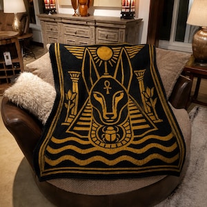 May include: A black and gold woven throw blanket featuring a central graphic of the Egyptian deity Anubis, an ankh symbol, a scarab beetle, stylized pillars, lotus flowers, and a sun disc, all framed by horizontal wave patterns at the bottom.