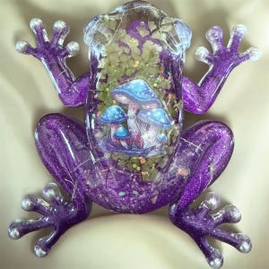 May include: A translucent purple resin frog figurine featuring a glittery finish and embedded green foliage. The center of the frog displays a detailed graphic illustration of several blue mushrooms with white stems, accented by small iridescent sparkles.