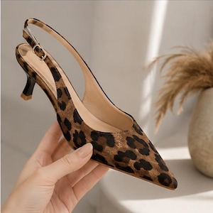 May include: A pointed-toe slingback shoe featuring a brown and black leopard print pattern. The shoe has a low kitten heel with a black base and a thin adjustable strap with a small gold-toned metal buckle at the side for a secure fit.