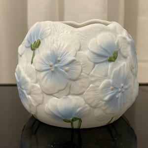 Op de afbeelding: Een witte keramische vaas met een ronde vorm, voorzien van reliëf bloemmotieven. De bloemen zijn beschilderd met zachte blauwe gradiënten op de bloemblaadjes, groene stengels en kleine gele hartjes, wat zorgt voor een textuurrijk, driedimensionaal uiterlijk.