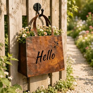 May include: A rust-coloured metal wall planter shaped like a handbag, featuring the word Hello cut out in black script and a small butterfly cutout. The planter is suspended from a black metal hook and contains small white and pink flowers.