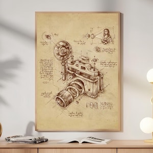 May include: A framed wall art print featuring a sepia-toned technical sketch of a vintage camera. The illustration includes detailed exploded-view components, a circular flash, and handwritten-style annotations, including a small portrait sketch.