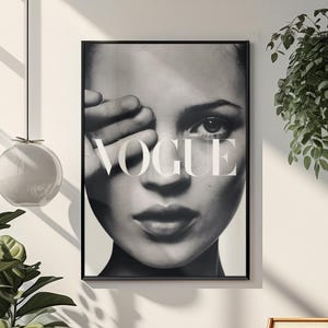 May include: A black and white framed wall art print featuring a close-up portrait of a face with one hand partially covering an eye. The word VOGUE is printed in large white capital letters across the center of the image, overlaying the facial features.