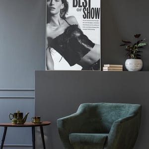 May include: A framed black and white poster featuring a person wearing a dark top with the text BEST OF SHOW. The poster is displayed above a dark green upholstered armchair with light wooden legs, next to a small wooden table with a green teapot.