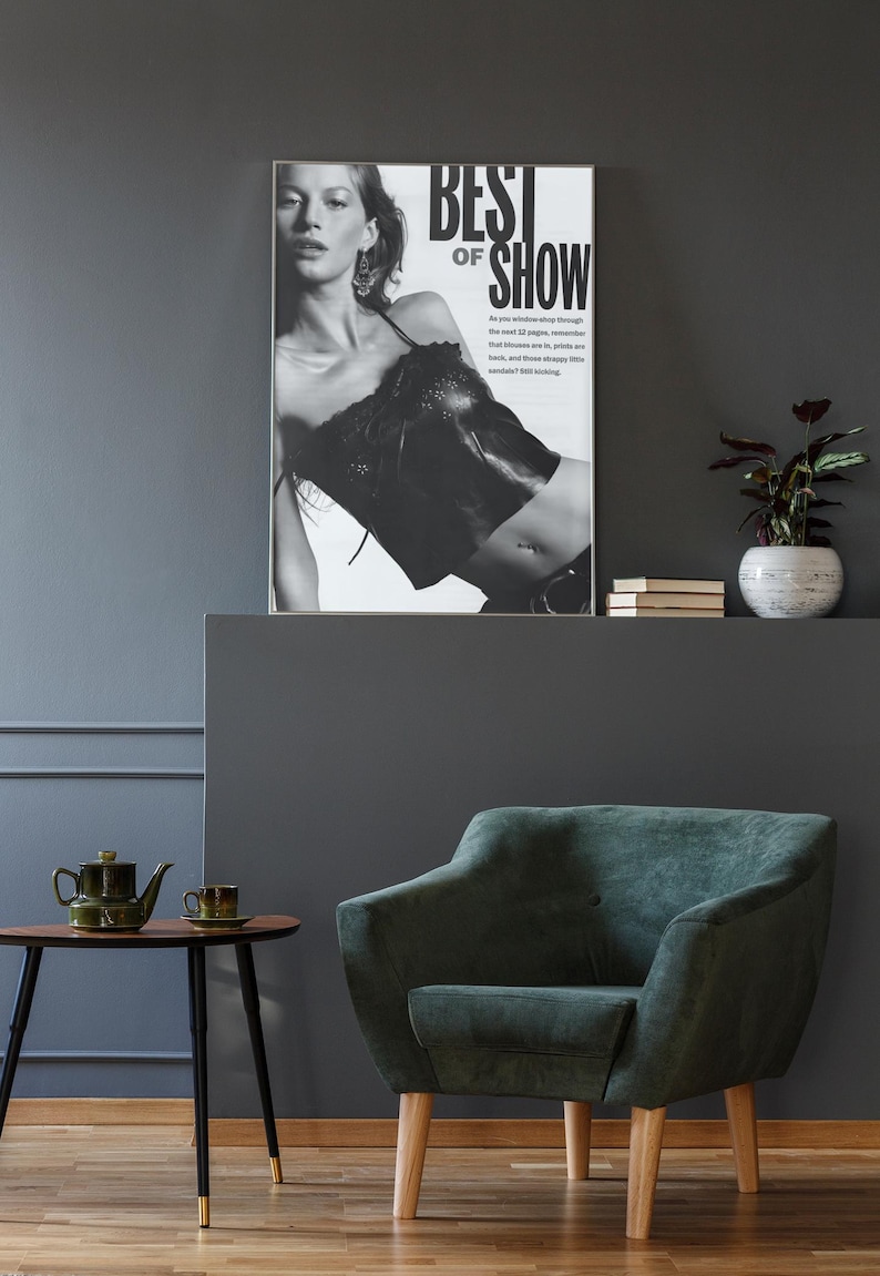 May include: A framed black and white poster featuring a person wearing a dark top with the text BEST OF SHOW. The poster is displayed above a dark green upholstered armchair with light wooden legs, next to a small wooden table with a green teapot.