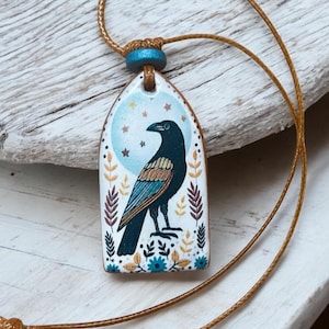 Può includere: Una collana con un ciondolo in ceramica che presenta un corvo nero con accenti blu e dorati in piedi di fronte a una luna bianca con stelle. Il ciondolo è circondato da un motivo floreale in tonalità di marrone, giallo e blu. La collana è realizzata con un cordoncino marrone.