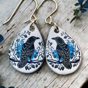 Puede incluir: Un par de pendientes en forma de lágrima con fondo blanco y una ilustración de cuervo azul y negro. Los pendientes tienen ganchos dorados.