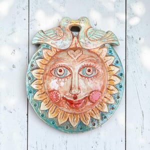 May include: A circular ceramic wall plaque featuring a sun face with a warm orange and yellow complexion, surrounded by a teal border. Two birds are perched at the top, touching beaks. The sun has detailed eyes, a nose, and a smiling mouth.