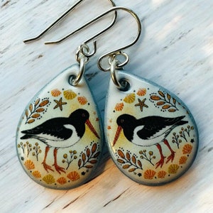 May include: A pair of teardrop-shaped ceramic earrings with silver hooks. Each earring features a black and white bird with an orange beak, surrounded by a floral design with seashells and starfish.
