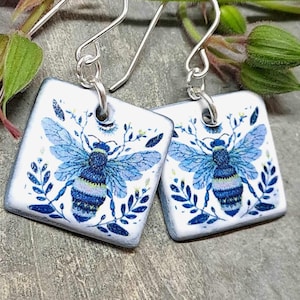 Puede incluir: Un par de pendientes de cerámica azul y blanca con un diseño de abeja. Los pendientes son de forma cuadrada y tienen ganchos de plata.