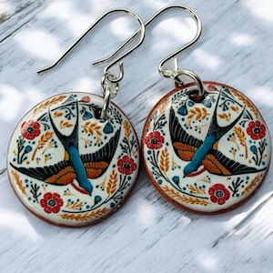 Puede incluir: Un par de pendientes redondos con un diseño de pájaro y flores. Cada pendiente presenta un fondo blanco con un pájaro negro, azul y naranja rodeado de flores rojas y follaje verde. Los pendientes tienen ganchos plateados.
