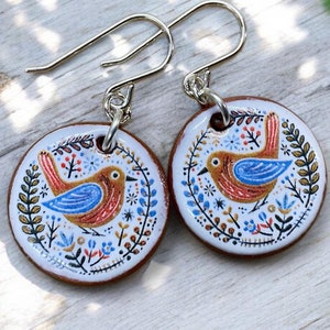 Puede incluir: Un par de pendientes de cerámica con fondo blanco y un diseño de pájaro colorido. El pájaro es rojo, azul y amarillo con un patrón floral a su alrededor. Los pendientes están unidos a ganchos de plata.