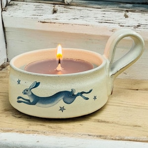 May include: A cream-coloured ceramic candle holder with a handle, featuring a blue hare and star design. A lit candle with dark purple wax sits inside. The holder is placed on a wooden surface, with a window in the background.