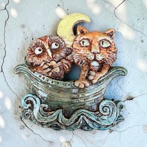 May include: A ceramic wall plaque featuring a brown owl and an orange cat sitting together in a small boat on stylised blue waves. A yellow crescent moon is positioned behind the animals. The piece has a glossy finish and textured details.