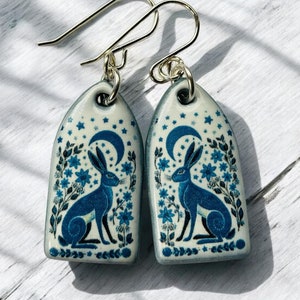 Può includere: Un paio di orecchini in ceramica con un design blu e bianco che presenta un coniglio e una luna crescente. Gli orecchini sono attaccati a ganci d'argento.