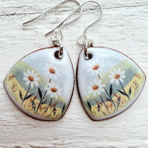 May include: A pair of handmade earrings featuring a daisy design. The earrings have a triangular shape with a white background and a painted scene of daisies in a field. The earrings have silver-colored ear wires.