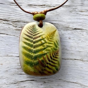 Puede incluir: Un collar con colgante rectangular con un diseño de helecho. El colgante tiene un fondo crema con una impresión detallada de helecho verde y marrón. El collar tiene un cordón marrón y una cuenta verde.