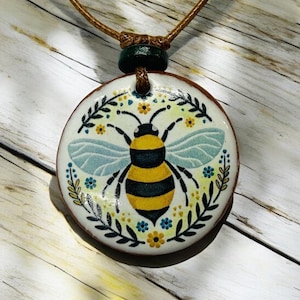 May include: A circular pendant necklace with a bee illustration. The bee is yellow and black, set against a background of blue and yellow floral and leaf patterns. The pendant is suspended from a brown cord with a green bead.