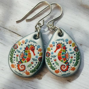 Puede incluir: Un par de pendientes de aro de plata con un dije de cerámica en forma de lágrima que presenta un diseño de caballito de mar colorido. El dije es blanco con un patrón floral azul, verde y naranja.