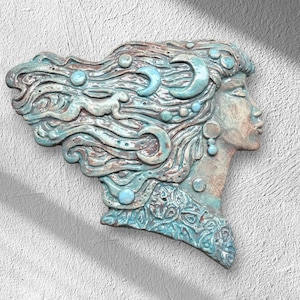 Pode incluir: Uma placa de parede de cerâmica apresentando o perfil lateral de um rosto com cabelos fluidos decorados com luas crescentes e formas circulares em relevo. A peça tem um acabamento desgastado em azul-petróleo e marrom com padrões espirais.