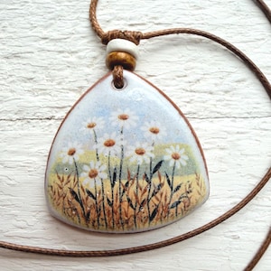 May include: A handcrafted pendant necklace featuring a painted enamel design of daisies in a field. The pendant is a light blue, triangular shape with a brown border, strung on a brown cord with a wooden bead.