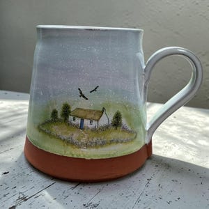 May include: A ceramic mug with a white handle and a terracotta-coloured base. The body features a hand-painted scene of a small white cottage with a blue door, surrounded by green grass, trees, and two birds flying in a soft, light-coloured sky.