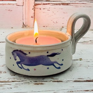 May include: White ceramic candle holder with a handle, featuring a purple horse design and a lit tea light candle inside.