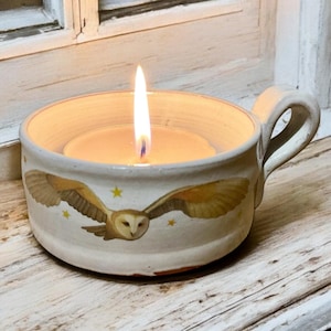 May include: A burning candle in a ceramic owl-themed cup. The cup is off-white with a handle and features a flying owl design with yellow stars. The candle flame is bright against the warm candle wax.