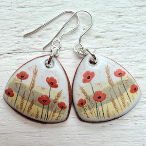 May include: A pair of handcrafted earrings featuring a floral design. Each earring has a white background with red poppies and wheat stalks. The earrings are teardrop-shaped with a brown border and silver ear wires.