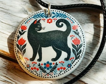 May include: A black cat with a curled tail is depicted on a round ceramic pendant. The pendant is white with a blue and red floral pattern. The pendant is attached to a black cord.