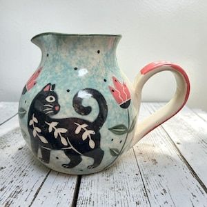 Può includere: Una brocca in ceramica con smalto azzurro maculato, caratterizzata da un gatto nero dipinto a mano con motivi a foglie bianche. La brocca è decorata con fiori rosa simili a tulipani e piccoli punti neri, e include un manico curvo con bordo rosa.