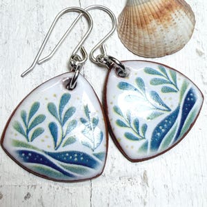 Puede incluir: Pendientes triangulares blancos con un diseño botánico en tonos azules y verdes. Los pendientes tienen un borde marrón y ganchos plateados. Una concha marina descansa en el fondo.