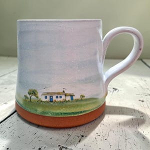 May include: Ceramic mug with a hand-painted landscape scene. The design features a small white house with blue doors and windows, set against a green field and a light blue sky. The base is a reddish-brown colour.