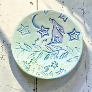 Puede incluir: Un plato redondo de cerámica con un diseño caprichoso. El plato presenta un fondo verde claro con un conejo azul, estrellas y una luna creciente. El borde tiene un patrón floral y de hojas en azul y verde.