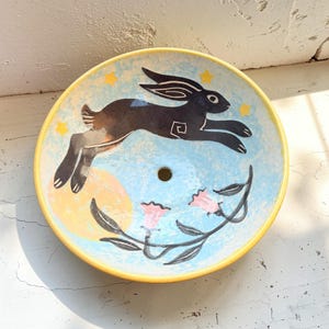 May include: A ceramic plate with a whimsical design. The plate features a black rabbit leaping across a light blue background with a yellow moon and stars. Pink flowers and a yellow rim complete the design.