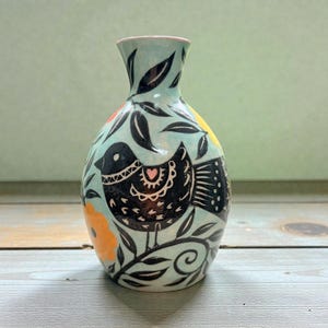 May include: A hand-painted ceramic vase with a light blue background, featuring a black bird design with a pink heart, surrounded by black leaves and orange and yellow flowers. The vase has a narrow neck and a rounded body.