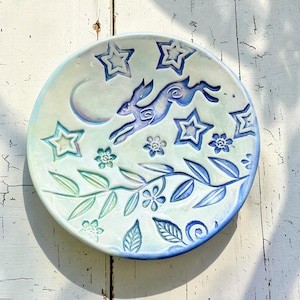 Puede incluir: Plato de cerámica con un diseño caprichoso. Presenta una liebre saltando, estrellas, una luna creciente y motivos florales y de hojas en tonos azules y verdes. El diámetro del plato es de unos 20 cm.